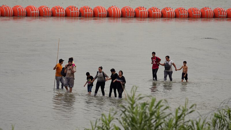 Migrantes que cruzaron el río Grande (Bravo) desde México caminan frente a enormes boyas...